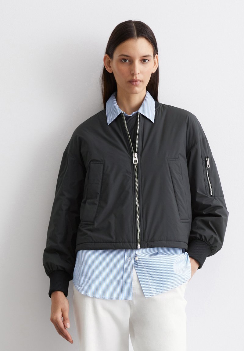 Schwarze Bomberjacke mit Reißverschluss, seitlichen Taschen und gerippten Bündchen, getragen über einem hellblauen gestreiften Hemd. Glatte Textur, cropped Design.