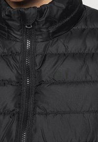 Černá prošívaná bunda s vysokým límcem, předním zipem a prošívanou strukturou s horizontálním prošitím.