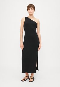 Vestido maxi negro de un solo hombro, hecho de un tejido suave, con una abertura lateral para mayor libertad de movimiento. Se lleva con sandalias negras planas.