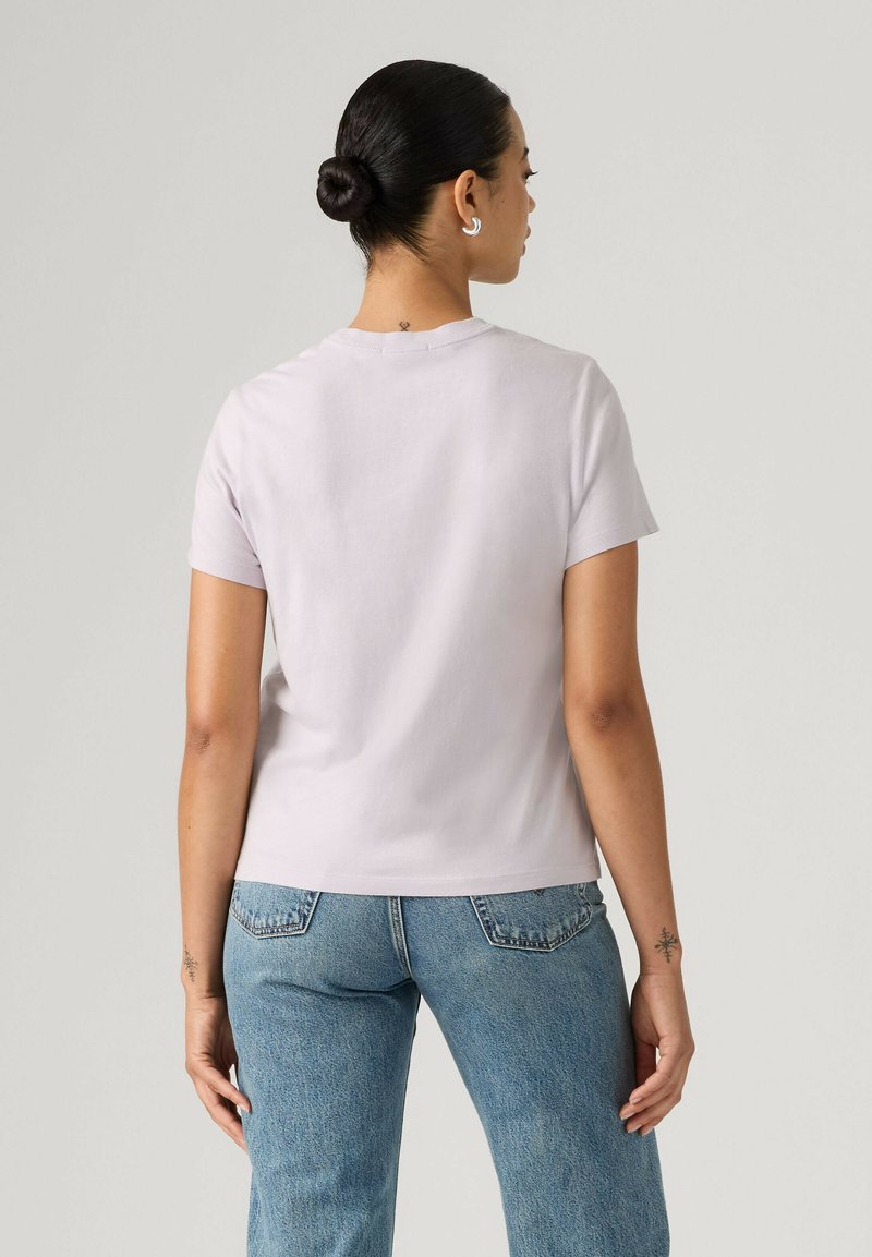 Femme avec les cheveux attachés en chignon, portant un t-shirt violet clair à manches courtes et un jean bleu, vue de dos sur un fond uni.