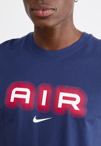 Μπλέ παραλλαγής βαμβακερό T-shirt με έντονο γραφικό "AIR" σε κόκκινο και λευκό χρώμα με γραμμικές λεπτομέρειες; περιλαμβάνει ένα μικρό λευκό λογότυπο Nike.