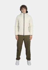 Giacca bianca imbottita con cerniere nere, chiusura frontale con zip e cappuccio. Pantaloni cargo verde oliva con tasche con zip. Il modello indossa sneakers bianche.
