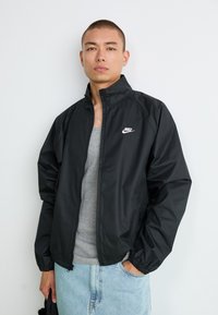 Ung mand iført en sort Nike zip-up jakke over en grå tanktop og lyseblå jeans, stående mod en ensfarvet hvid baggrund.