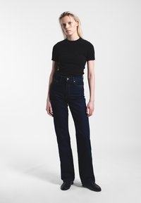 Jonge vrouw met blond haar die een zwart kort mouwtopje, donkere hooggetailleerde jeans en zwarte schoenen draagt, staand tegen een effen lichte achtergrond.