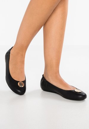 Jambes portant des ballerines en cuir noir avec un emblème logo circulaire doré sur les orteils, debout sur un sol blanc lisse.