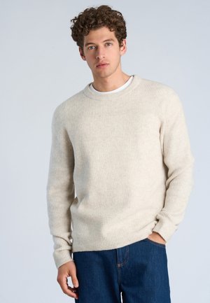 Ung mand med krøllet hår iført en cremefarvet ribstrikket sweater og blå jeans, stående mod en ensfarvet lysegrå baggrund.
