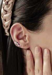 Cuff auricolare in argento con molte piccole pietre trasparenti disposte in un design floreale, indossato su un orecchio, con un dito che sfiora delicatamente la guancia.