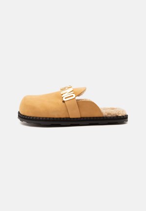 Brązowe chodaki slip-on z czarną podeszwą, podszyte futrem z baranka, z szerokim paskiem na górze z dużymi złotymi literami.
