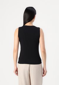 Mujer con cabello largo y oscuro, vistiendo una blusa sin mangas negra y pantalones de un beige claro, de pie de espaldas contra un fondo blanco liso.