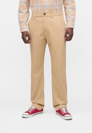 Άνδρας που φοράει μπεζ παντελόνια με ίσια γραμμή, παστέλ καρό πουκάμισο με κουμπιά και κόκκινα sneakers χαμηλού πάτου με λευκά κορδόνια.