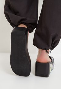 Czarne sandaly na platformie z teksturowaną podeszwą i kolorowymi wzorami w stylu graffiti na cholewce. Obcas jest masywny i kwadratowy.