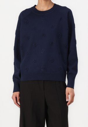 Personne portant un pull bleu marine avec des motifs géométriques subtils et un pantalon noir à jambes larges sur un fond blanc uni.