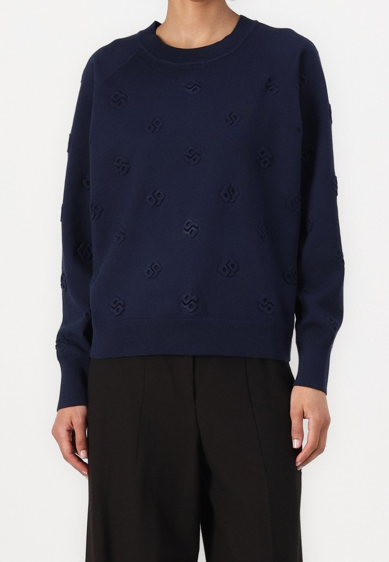 Personne portant un pull bleu marine avec des motifs géométriques subtils et un pantalon noir à jambes larges sur un fond blanc uni.
