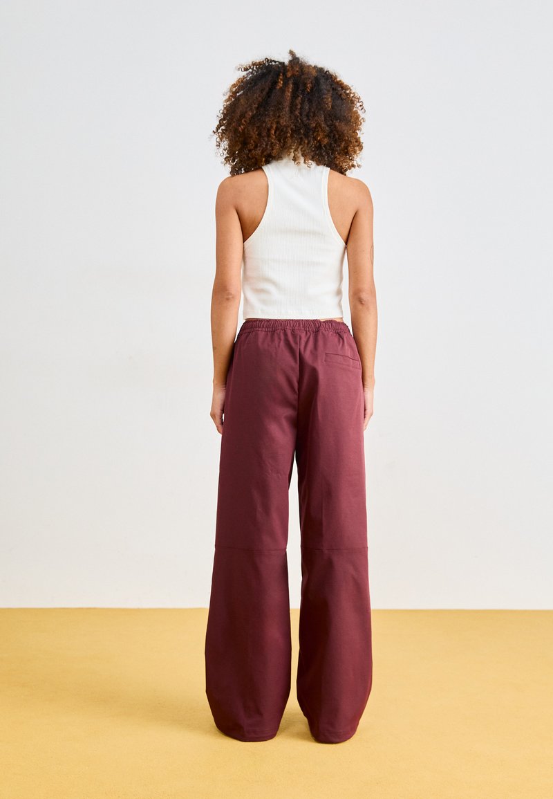 Une femme aux cheveux bouclés se tient dos à nous, portant un haut sans manches blanc et un pantalon large maroon ample sur un sol jaune.