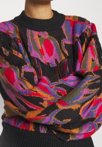 Pull en maille multicolore présentant des motifs vibrants en rouge, violet, orange et vert, avec un col côtelé noir et des détails à franges.