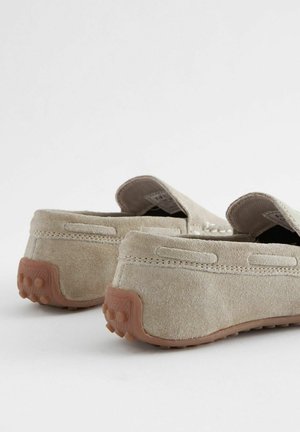 Pantofi slip-on din suede bej, cu o talpă din cauciuc texturată și accente cusute. Partea din spate are o formă rotundă cu puncte mici de prindere.