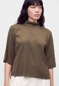 Jeune femme aux longs cheveux foncés porte un haut vert olive à col montant et manches courtes ainsi qu'un pantalon blanc sur un fond uni.