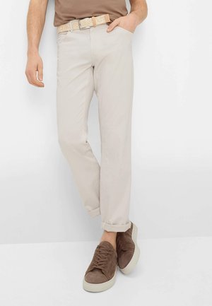 Mand iført beige bukser med opkrogne ben, et struktureret beige bælte, brune casual sneakers og en brun skjorte, med den ene hånd i lommen.