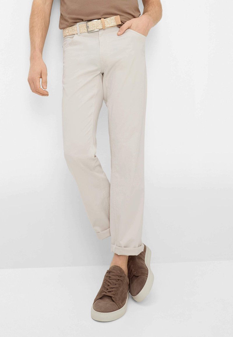 Mann trägt beige Hose mit hochgekrempelten Säumen, einen strukturierten beigen Gürtel, braune Freizeitsneaker und ein braunes Hemd, eine Hand in der Tasche.