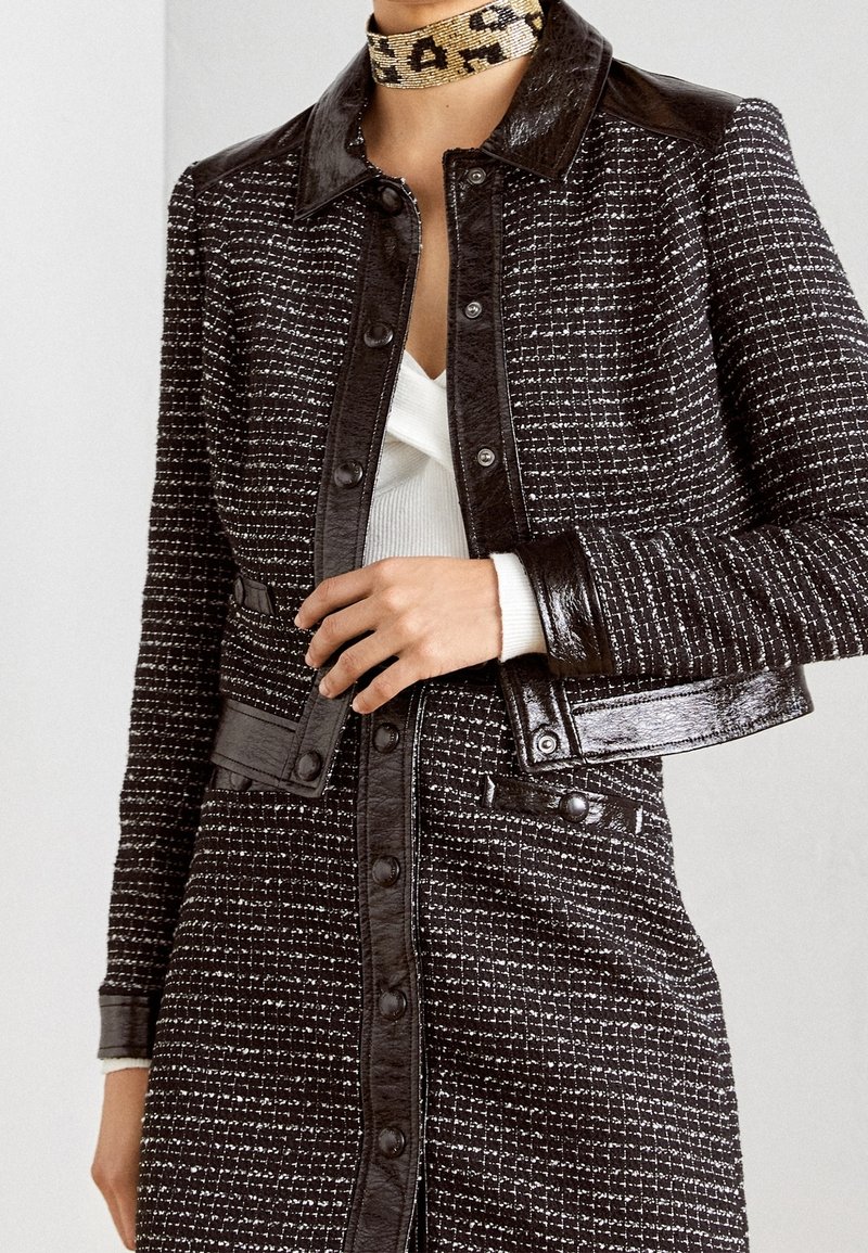 Femme portant une veste en tweed noir et blanc avec des finitions en cuir et des boutons, par-dessus un haut blanc, et un collier ras-du-cou imprimé léopard.