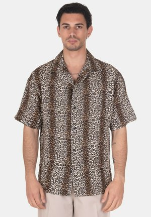 Uomo che indossa una camicia a maniche corte con fantasia animalier marrone e nera, in piedi davanti a uno sfondo chiaro e uniforme.