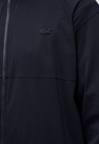 Veste noire zippée avec logo crocodile texturé sur la poitrine gauche et détail de couture sur le devant et la manche.