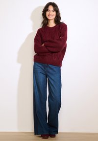 Bordowy sweter w warkocze z opuszczonymi ramionami, zestawiony z szerokimi dżinsami o luźnych nogawkach. Miękka tekstura z prążkowanym wykończeniem na dole i mankietach.