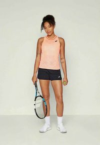 Sieviete persiku krāsas bezpiedurkņu topā un melnā šortā tur tennisu raketi vienā rokā un tenisa bumbu otrā, stāvot pret vienkāršu fona.