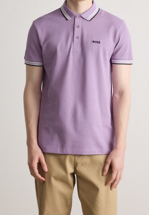 Mand iført lyse lilla kortærmet poloshirt med hvid og sort kant, "BOSS" logo og beige bukser, stående mod ensfarvet baggrund.