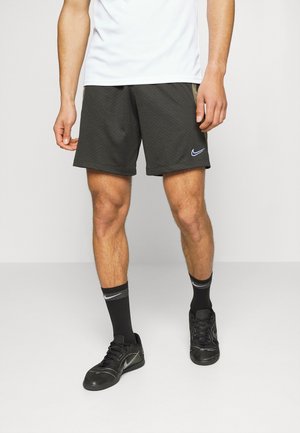 Mężczyzna ubrany w czarne spodenki Nike, czarne skarpety Nike i czarne buty sportowe Nike, stojący na jednolitym jasnym tle.