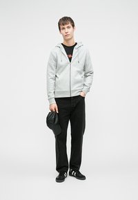 Hanorac gri cu fermoar și șnur, tricou negru, pantaloni negri și adidași negri; ține o șapcă neagră. Design simplu și croială lejeră.