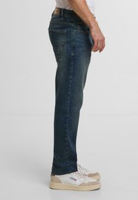 Jeans in denim blu scuro con vestibilità slim, caratterizzati da cinque tasche, un girovita standard e un leggero sbiadimento. Abbinati a sneakers bianche e color sabbia.