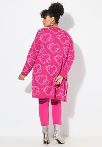 Růžový svetr s bílým vzorem srdce, oversize střih, dlouhé rukávy, doplněný o růžové kalhoty a stříbrné vysoké boty na podpatku.