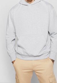 Felpa grigia realizzata in tessuto morbido, con tasca a marsupio e polsini a costina. Il design include un cappuccio a doppio strato. Abbinata a pantaloni beige.
