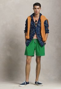 Model îmbrăcat cu o cămașă albastră cu model, un pulover portocaliu, un tricou cu dungi, pantaloni scurți verzi și adidași blue. Outfit simplu și casual.