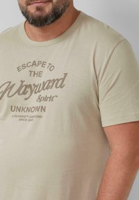 Beige t-shirt met korte mouwen van zacht katoen, met een bedrukte opdruk van "Escape to the Wayward Spirit" en "UNKNOWN" in bruin.