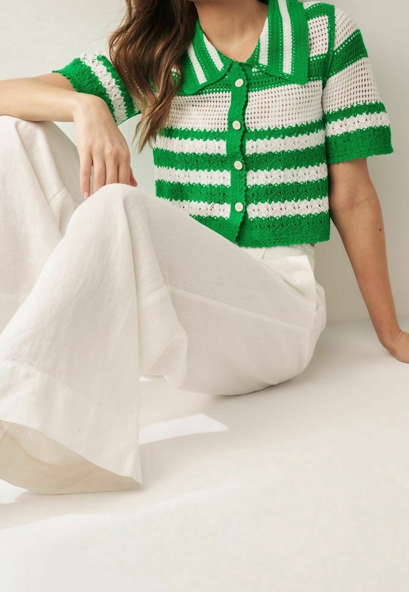 Femme assise portant un pantalon blanc à jambes larges et un top court en crochet rayé vert et blanc, manches courtes, boutonné avec col.