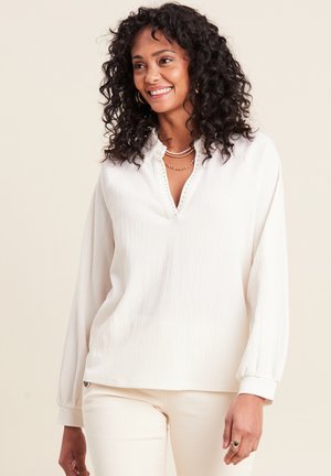 Weißer Pullover mit langen Ärmeln aus strukturiertem Stoff, V-Ausschnitt mit Knopfdetails und eng anliegenden Bündchen. Das Model trägt cremefarbene Hosen.