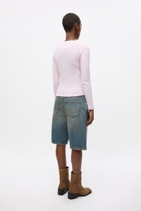 Maglietta a maniche lunghe rosa abbinata a pantaloni Bermuda in denim blu, con orlo sfrangiato. Stivali alla caviglia marroni completano l'outfit.
