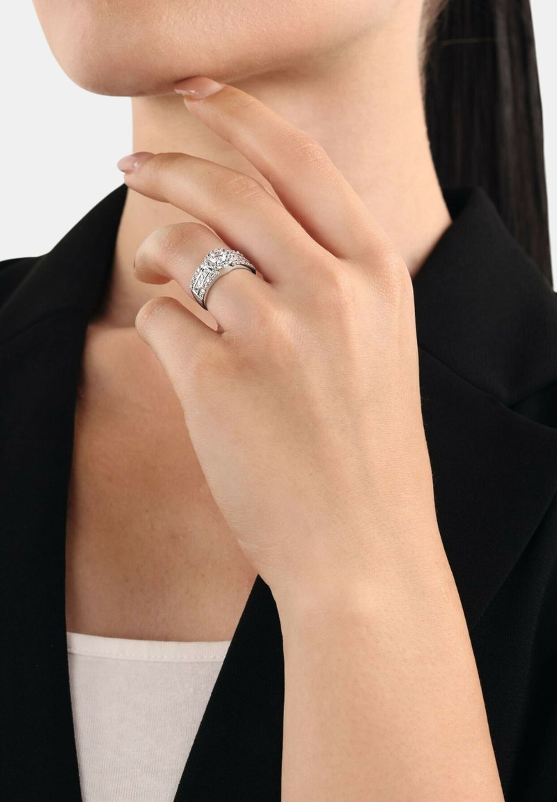 Anello in argento con dettagli intricati, dotato di una pietra centrale e una fascia texturizzata, indossato su una mano vicino al mento.