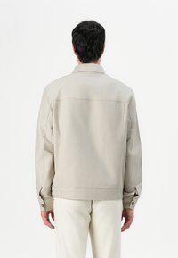 Beige canvas jas met lange mouwen, een kraag en knoopmanchetten. Heeft een relaxte pasvorm en een rechte rugontwerp.
