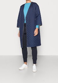 Cappotto lungo trapuntato navy con una vestibilità morbida, maniche ampie e apertura frontale. Indossato su un maglione turchese e pantaloni scuri, abbinato a sneaker bianche.