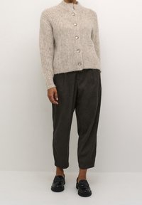 Cárdigan de punto color beige claro con un cuello alto y cinco botones plateados, combinado con pantalones sueltos gris oscuro y mocasines negros.