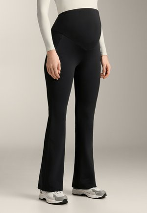 Pantaloni maternità neri realizzati in tessuto elasticizzato, con vita alta, design aderente e gambe svasate, abbinati a sneakers bianche.