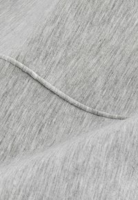 Hellgrauer Stoff mit subtiler melierter Textur, der gestickte Details und sanfte Falten zeigt. Weiches, dehnbares Material mit glatter Oberfläche.