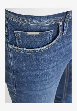 Primer plano del bolsillo delantero de unos vaqueros de mezclilla azul con un pequeño bolsillo para monedas que muestra una etiqueta de metal de la marca y costuras visibles en color naranja.