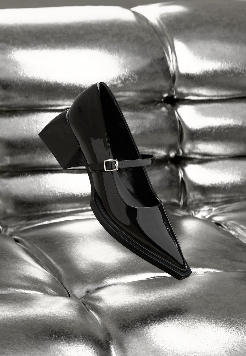 Chaussure à talon haut en cuir verni noir avec un bout pointu, une bride à la cheville et un talon carré. Présentée sur un fond argenté brillant.