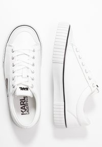 Weiße Sneakers mit einem glatten Lederobermaterial, runder Zehenpartie, Schnürdesign und Gummisohle mit ausgeprägten Streifen und metallischem Akzent.