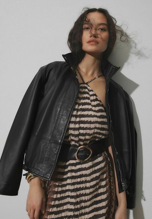 Kobieta z rozpuszczonymi kręconymi włosami, ubrana w czarną skórzaną kurtkę na wzorzystej plisowanej sukience z szerokim paskiem, stoi na tle gładkiej ściany.