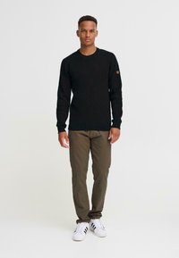 Pulover negru tricotat cu mâneci lungi, guler rotund, tiv striat și un mic logo pe mâneca stângă. Asortat cu pantaloni măslin și adidași albi.